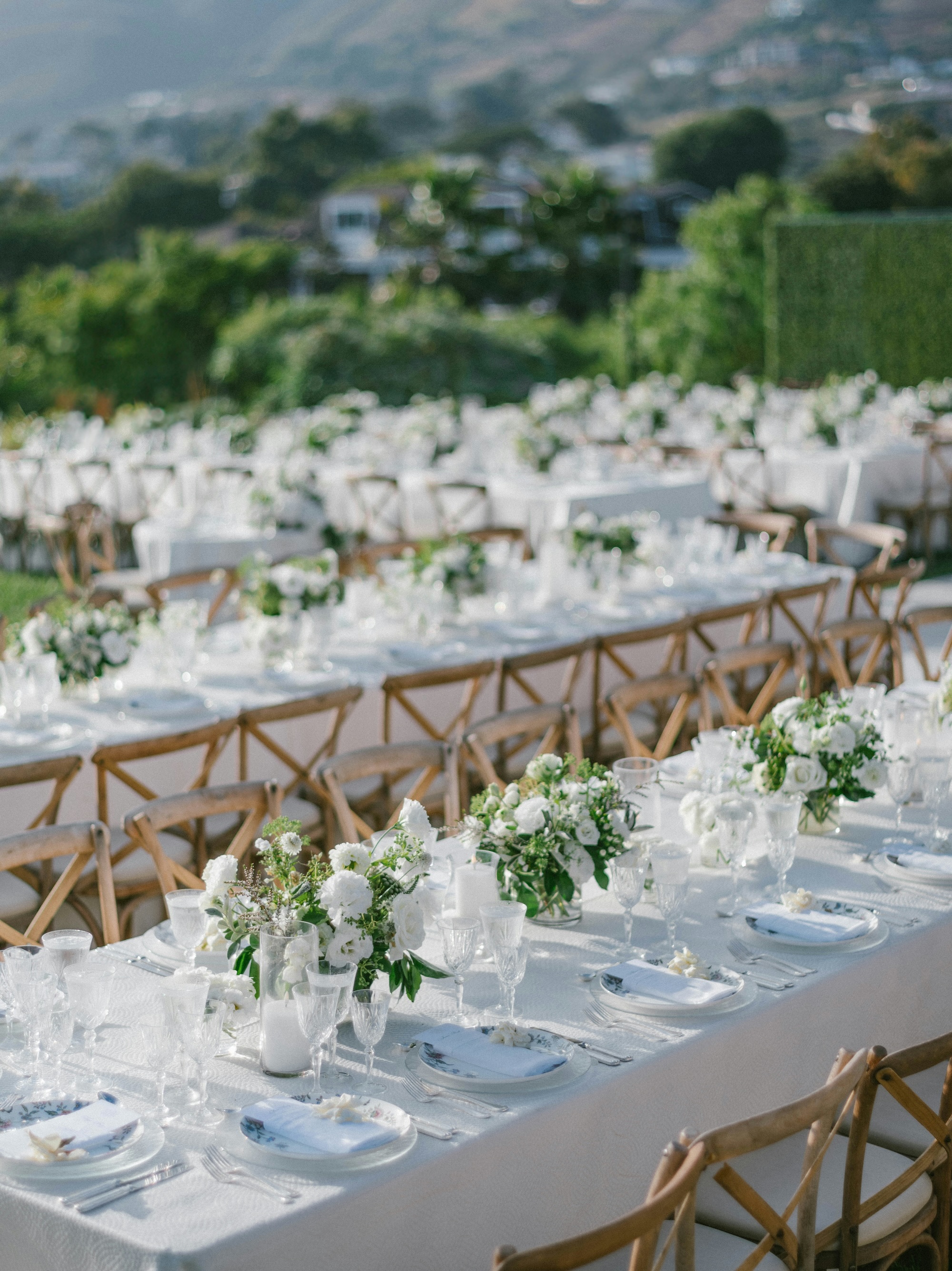 Long outdoor banquet tables with white tablecloths, floral centerpieces, crystal glassware, white plates, and wooden chairs set up for an elegant event.