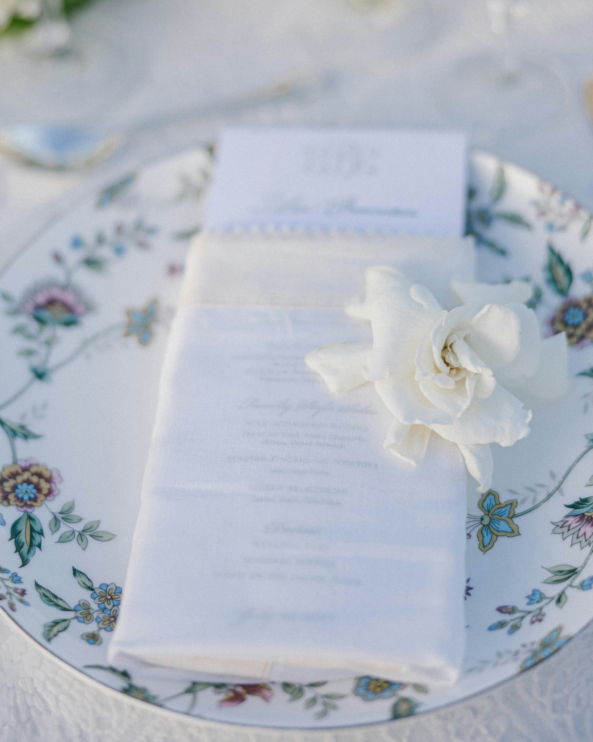 White cloth napkin with a white flower and a menu card placed on a decorative floral plate.