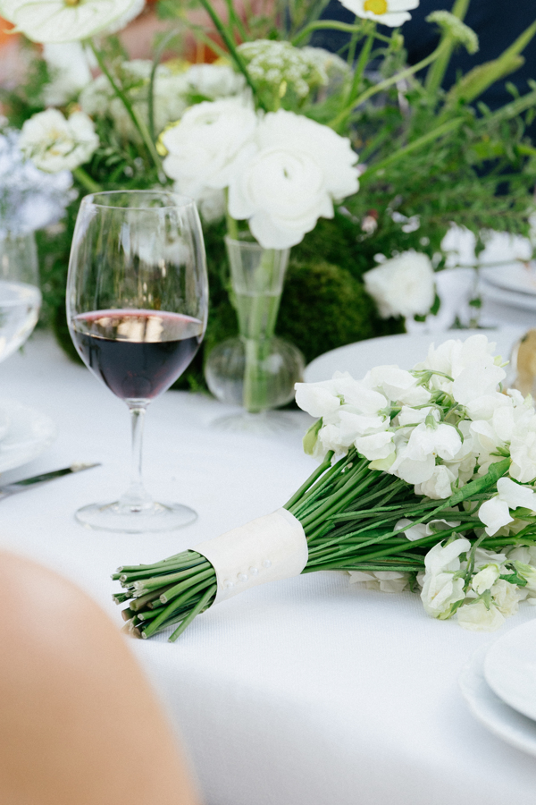 Close-up of a bouquet of white flowers and a glass of red wine on a white tablecloth with floral centerpiece in the background.