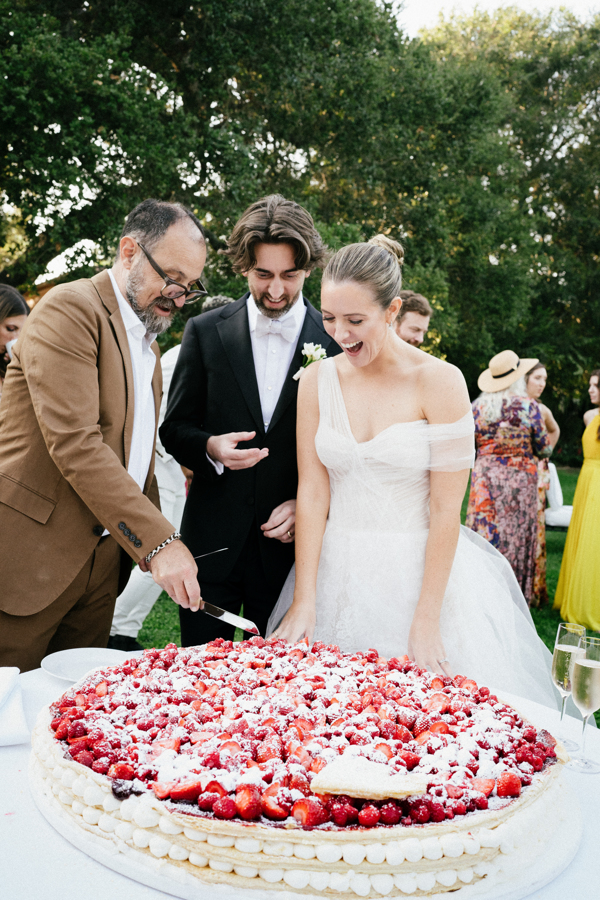 Bride and groom smiling as a man in a brown suit cuts a large fruit-topped wedding cake outdoors.