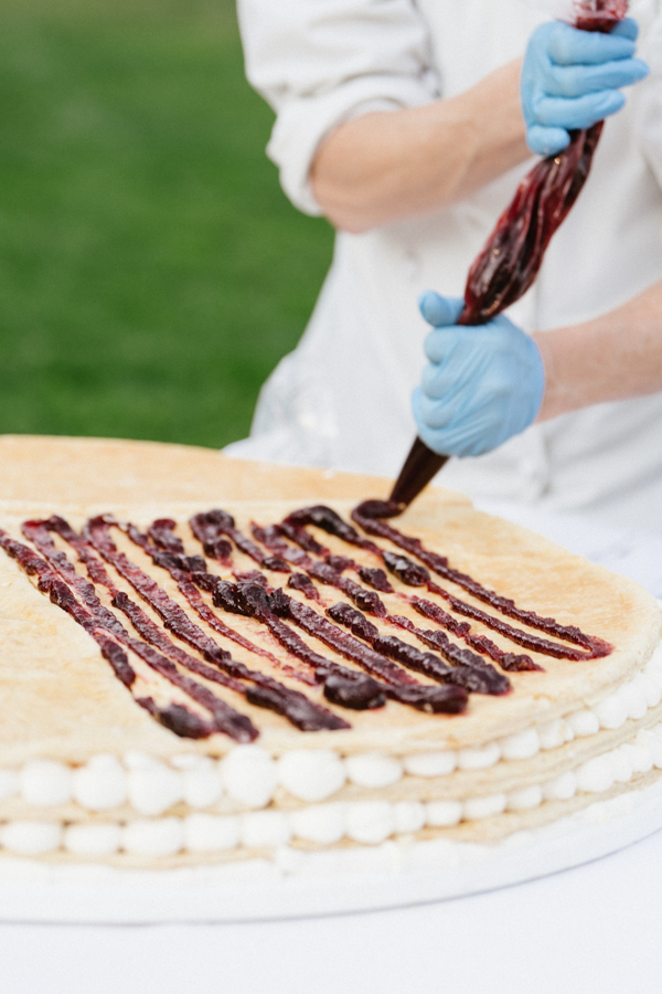Person wearing blue gloves decorating a layered cake with dark red fruit jam using a piping bag.