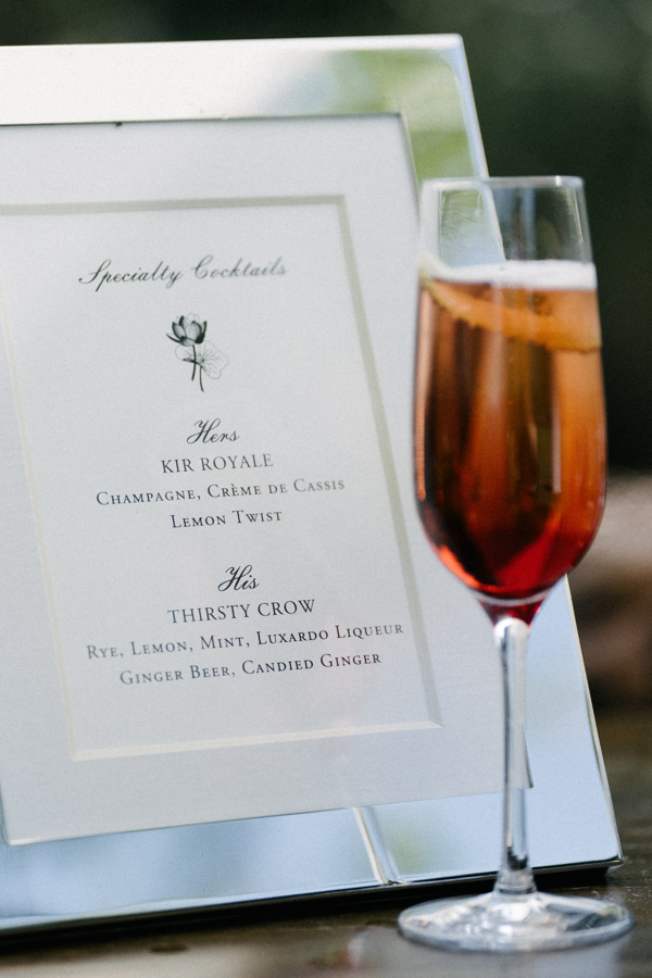 Specialty cocktails menu with 'Hers' Kir Royale made with champagne, crème de cassis, lemon twist and 'His' Thirsty Crow made with rye, lemon, mint, Luxardo liqueur, ginger beer, and candied ginger, next to a tall glass of a reddish cocktail.