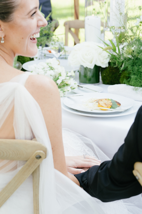 Smiling bride in a white dress holding hands with a person in a black suit at a wedding dinner table decorated with white flowers and greenery.