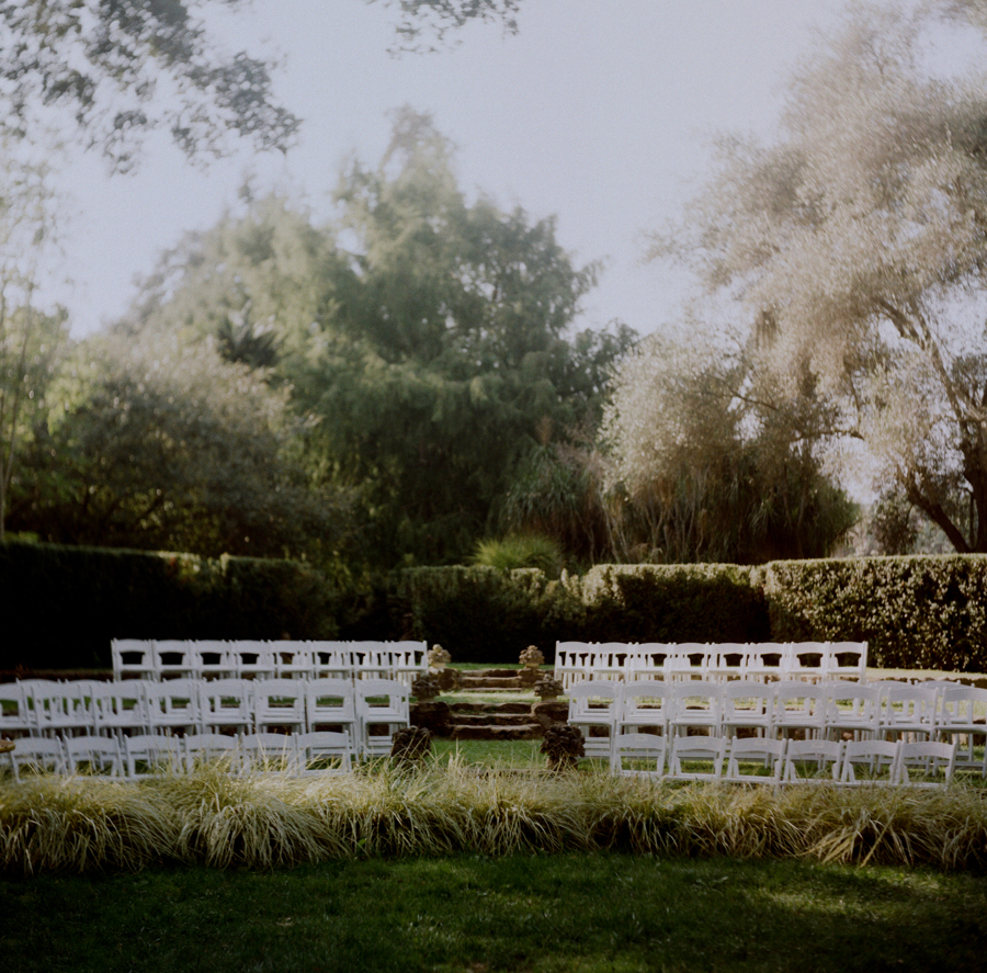 Outdoor ceremony setup with rows of white chairs facing a small stone staircase surrounded by greenery and trees.