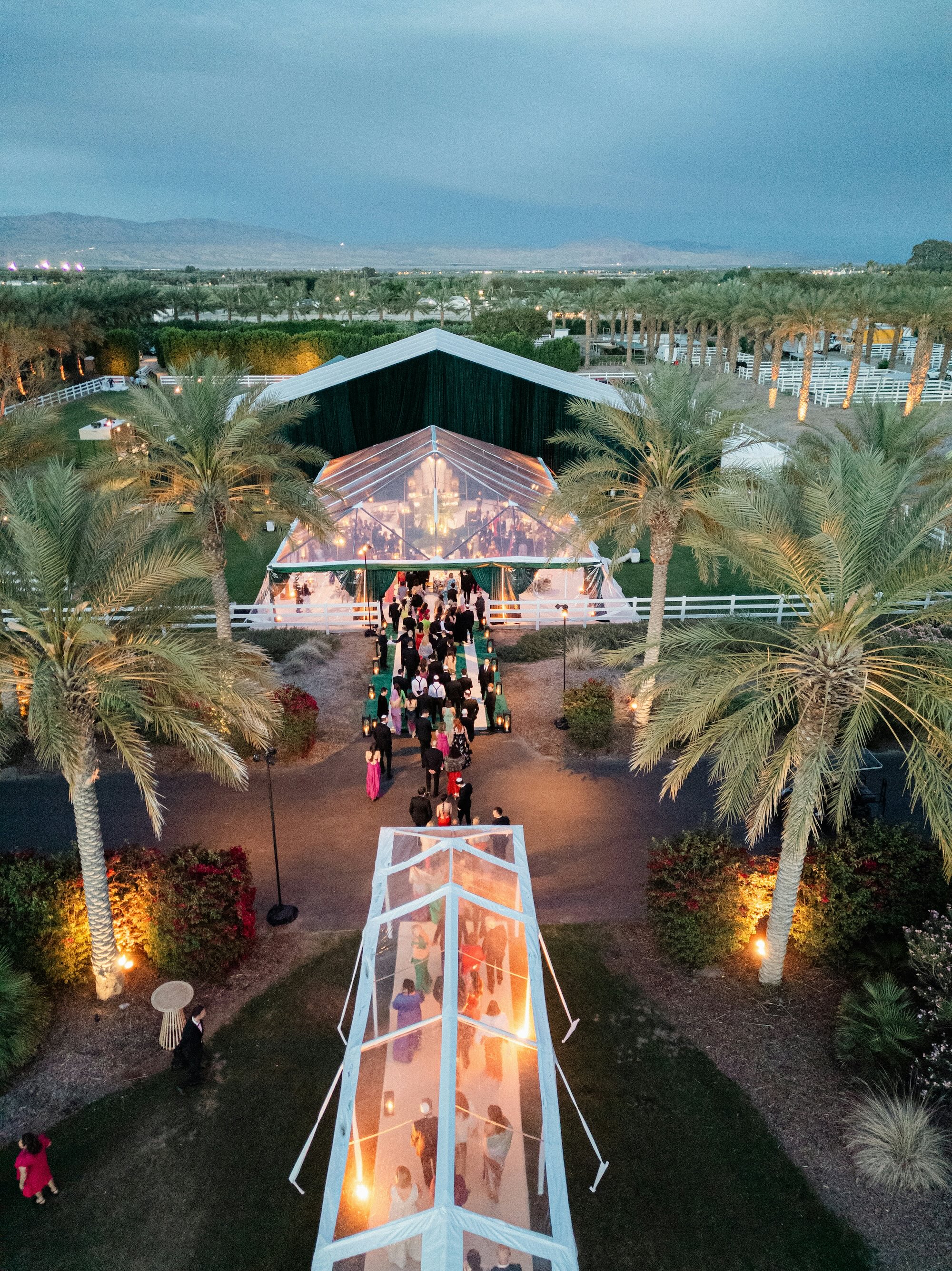 Aerial view of guests walking through a clear tented pathway lined with palm trees and ambient lighting towards a large, illuminated event tent set in a landscaped outdoor area at dusk.