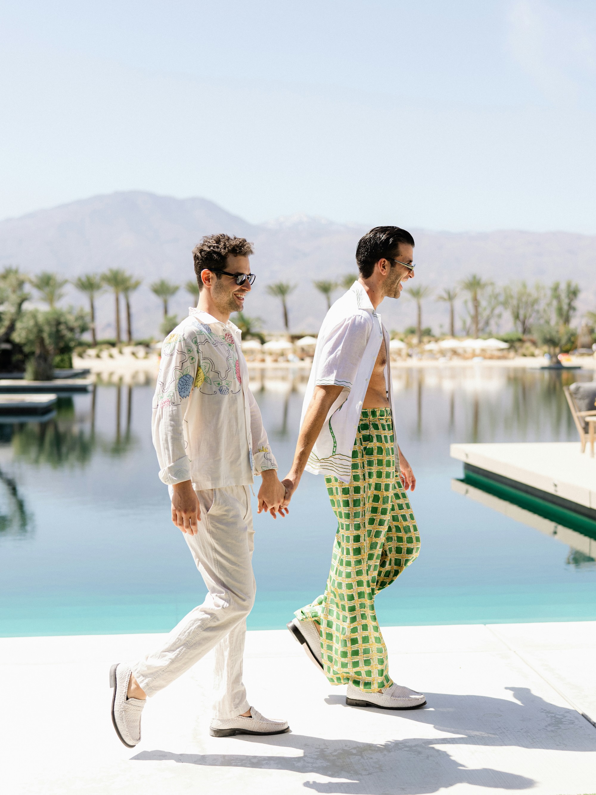 Two men holding hands and walking beside a pool with mountains and palm trees in the background.