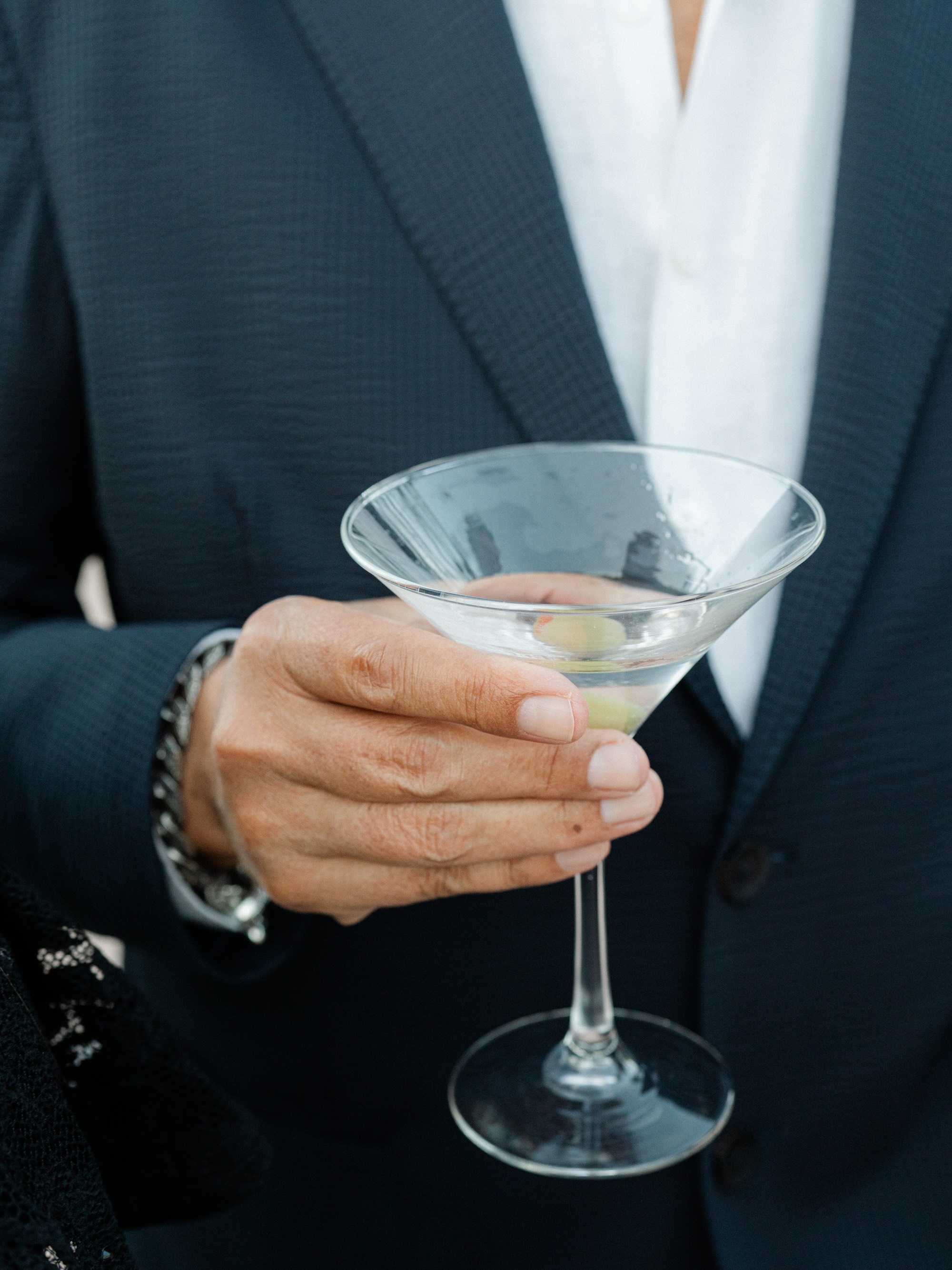 Close-up of a person in a dark suit holding a martini glass with two olives.