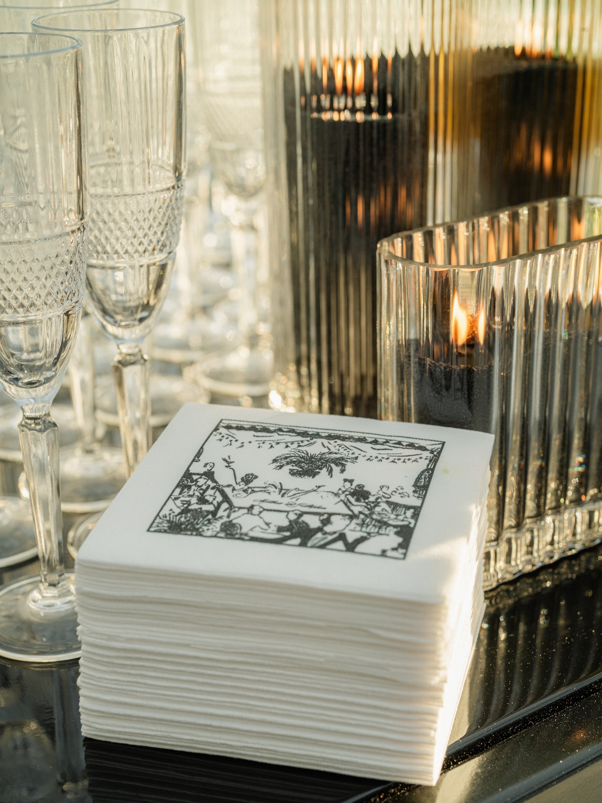 Stack of white napkins with a black printed design next to lit candles in ribbed glass holders and empty crystal champagne flutes.