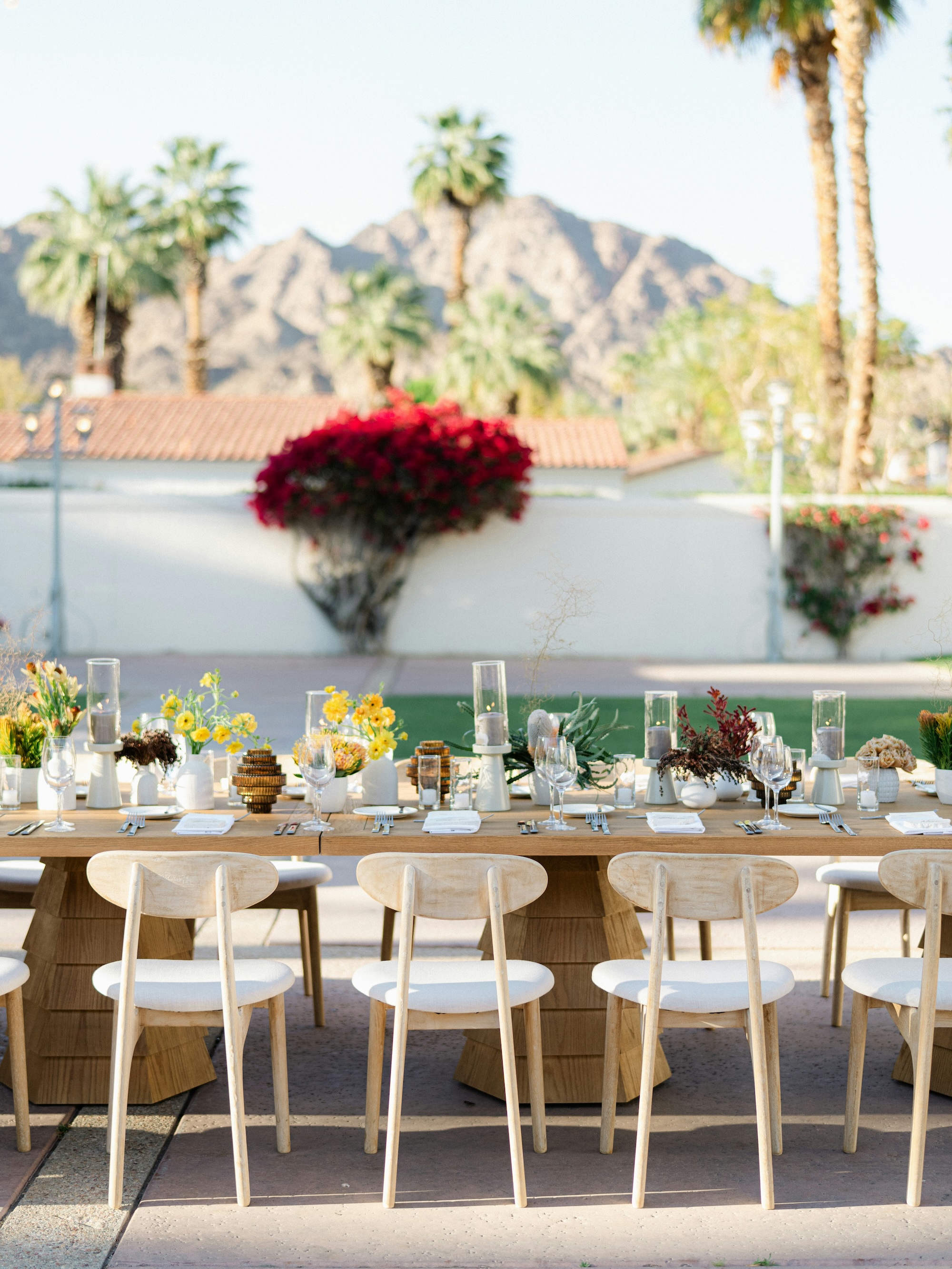 Outdoor wooden dining table set for event with flower centerpieces, candle holders, and white cushioned chairs, mountains in background.