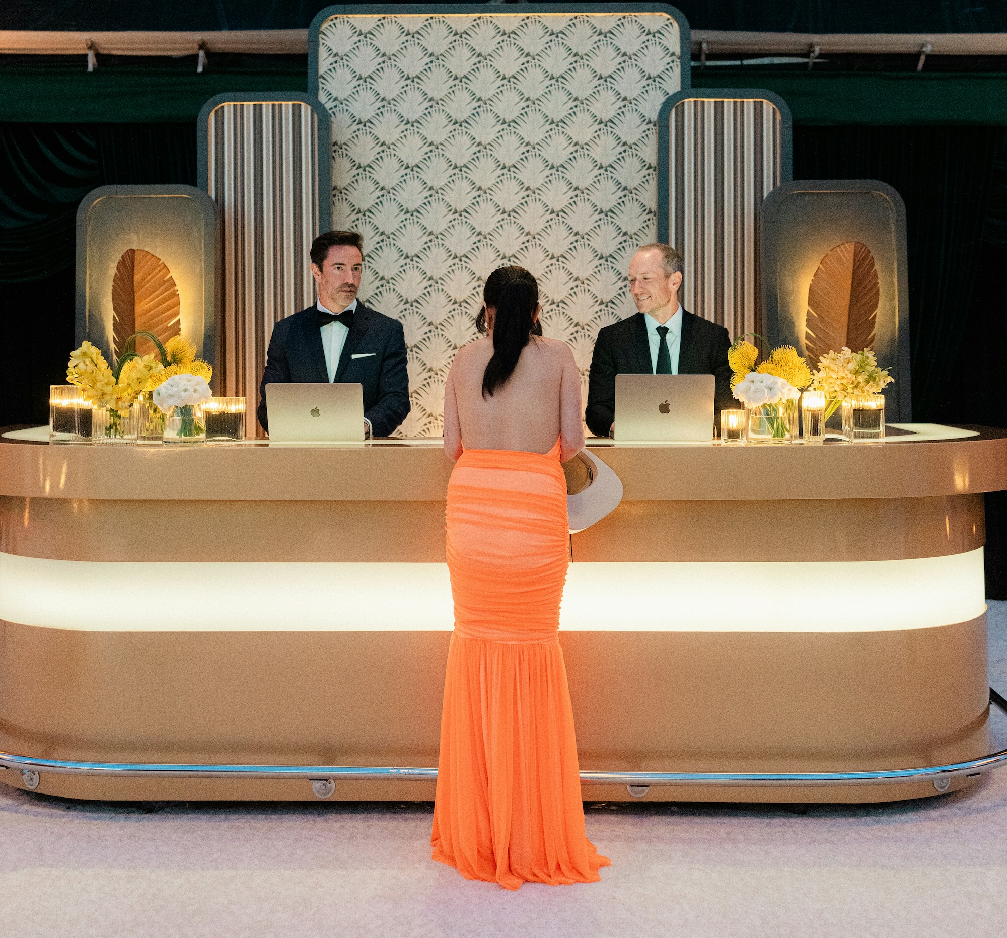 Woman in a bright orange backless gown stands at a modern reception desk with two men in tuxedos working on laptops behind it.