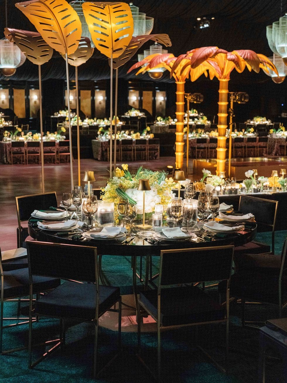 Elegant round dining table set with glassware, white napkins, candles, and floral centerpiece under golden leaf-shaped decorations in a dimly lit banquet hall.