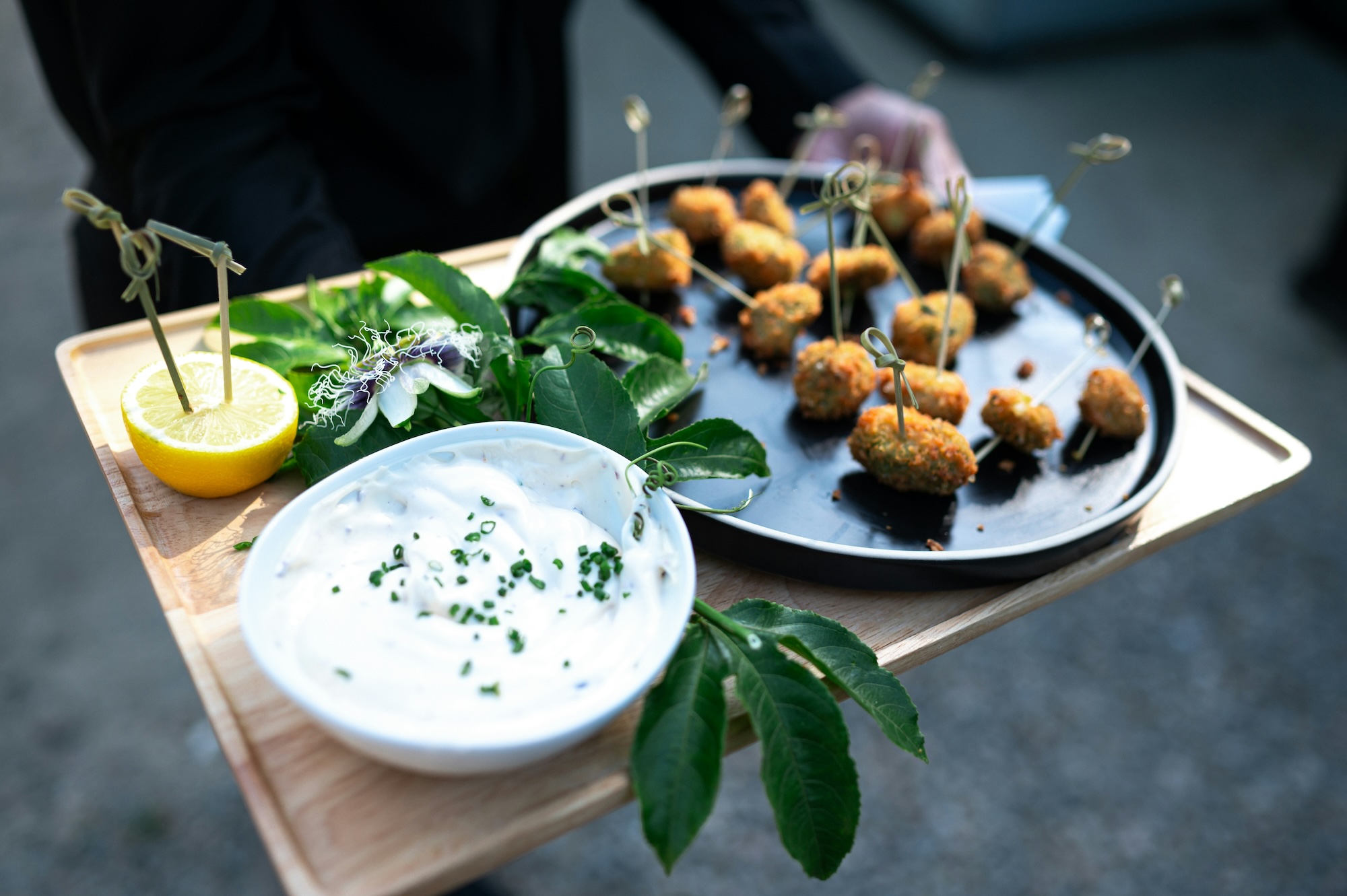 Tray with bite-sized fried appetizers on skewers served with creamy dip garnished with chives, lemon half, and green leaves.