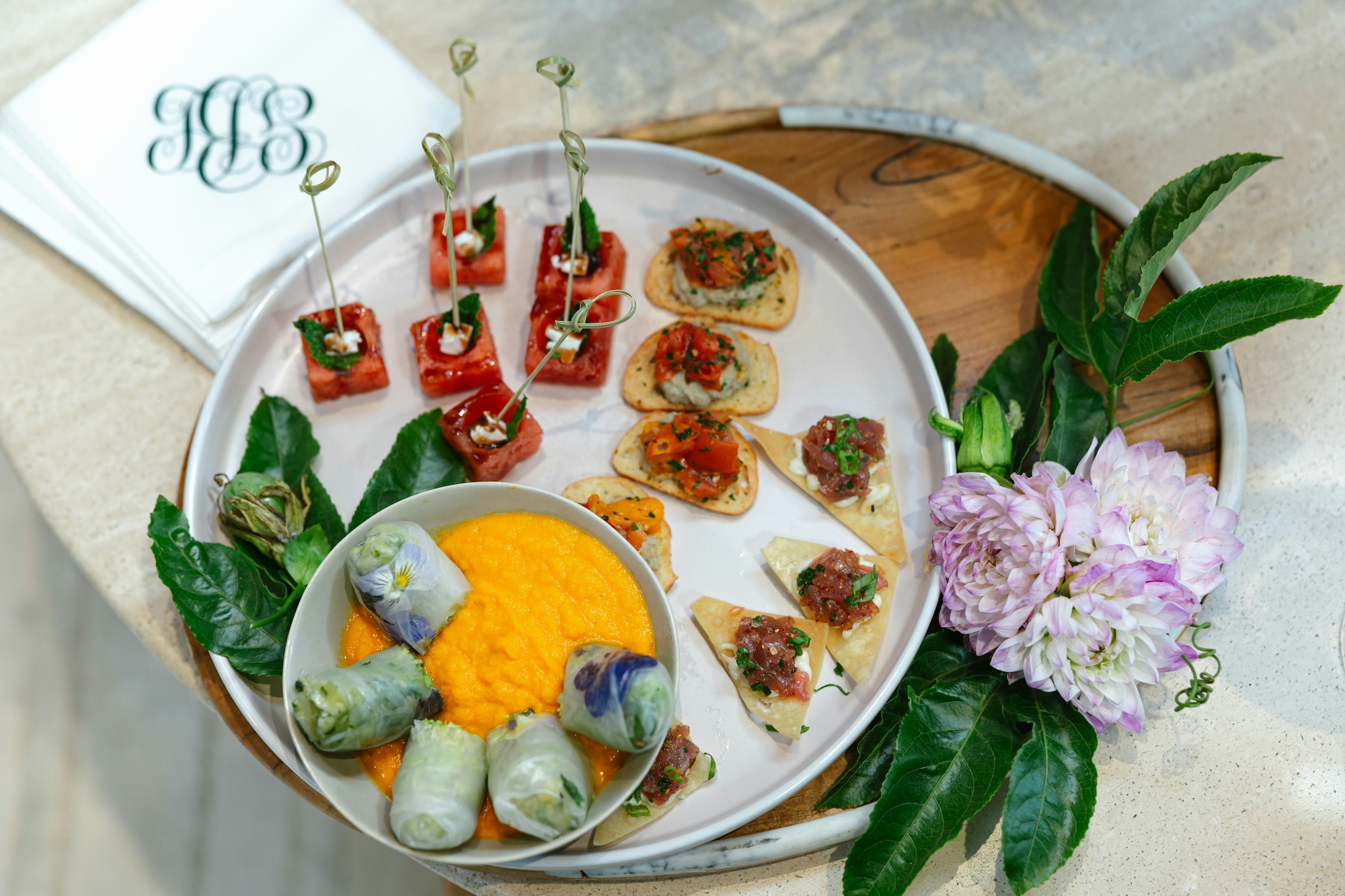 White round plate with assorted appetizers including watermelon cubes with herbs, small toast with diced tomatoes, triangular crackers with tuna tartare, and fresh spring rolls around a bowl of orange dipping sauce, garnished with green leaves and light purple flowers.