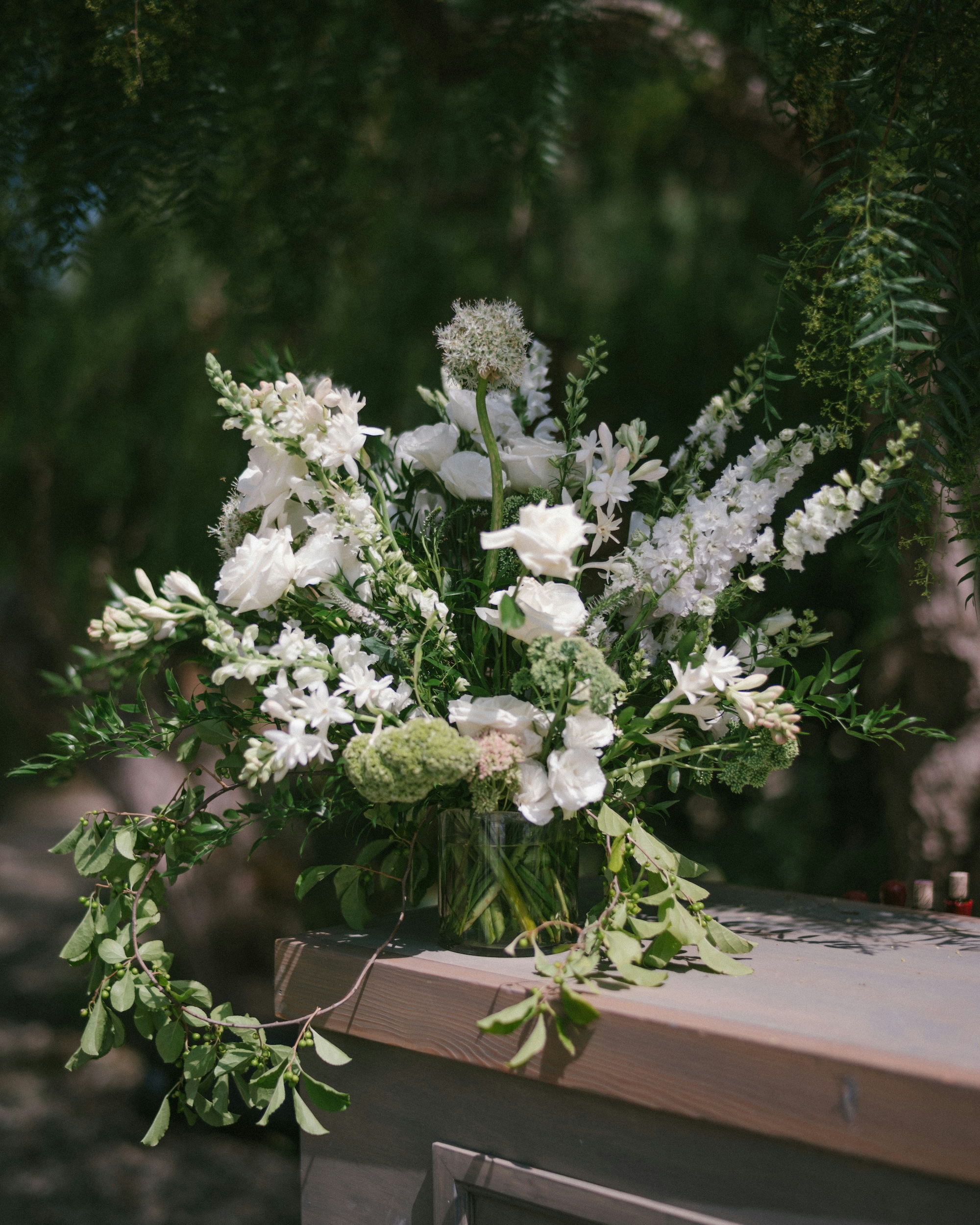 Elegant bouquet of white flowers and green foliage in a glass vase on a wooden surface outdoors.