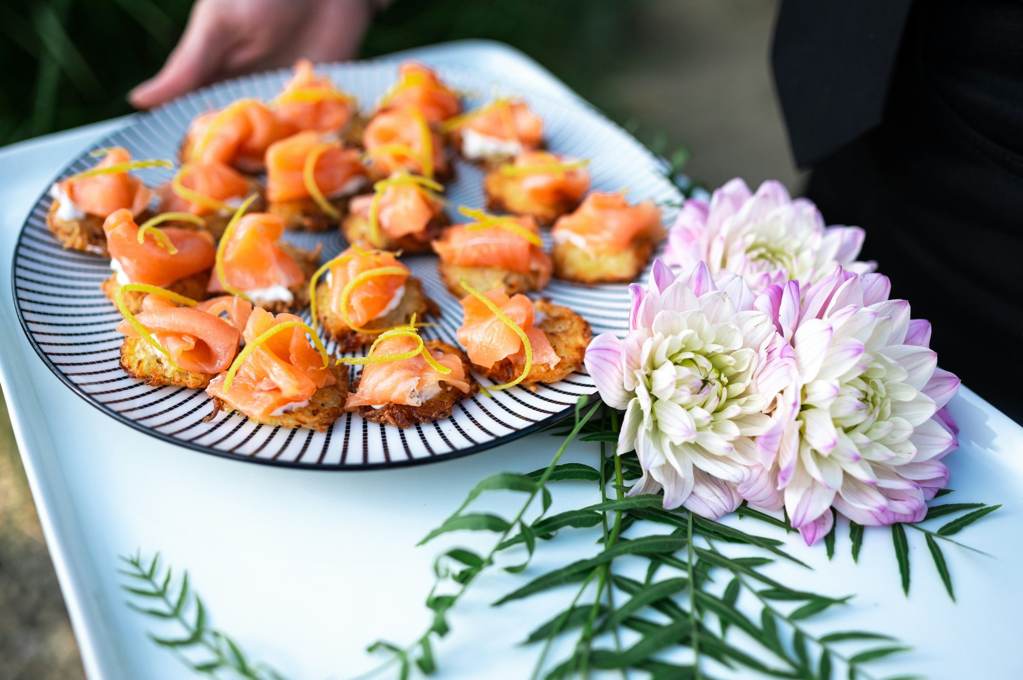 Plate of smoked salmon bites on crispy bases garnished with lemon zest, held on a tray decorated with three white and pink flowers with green leaves.