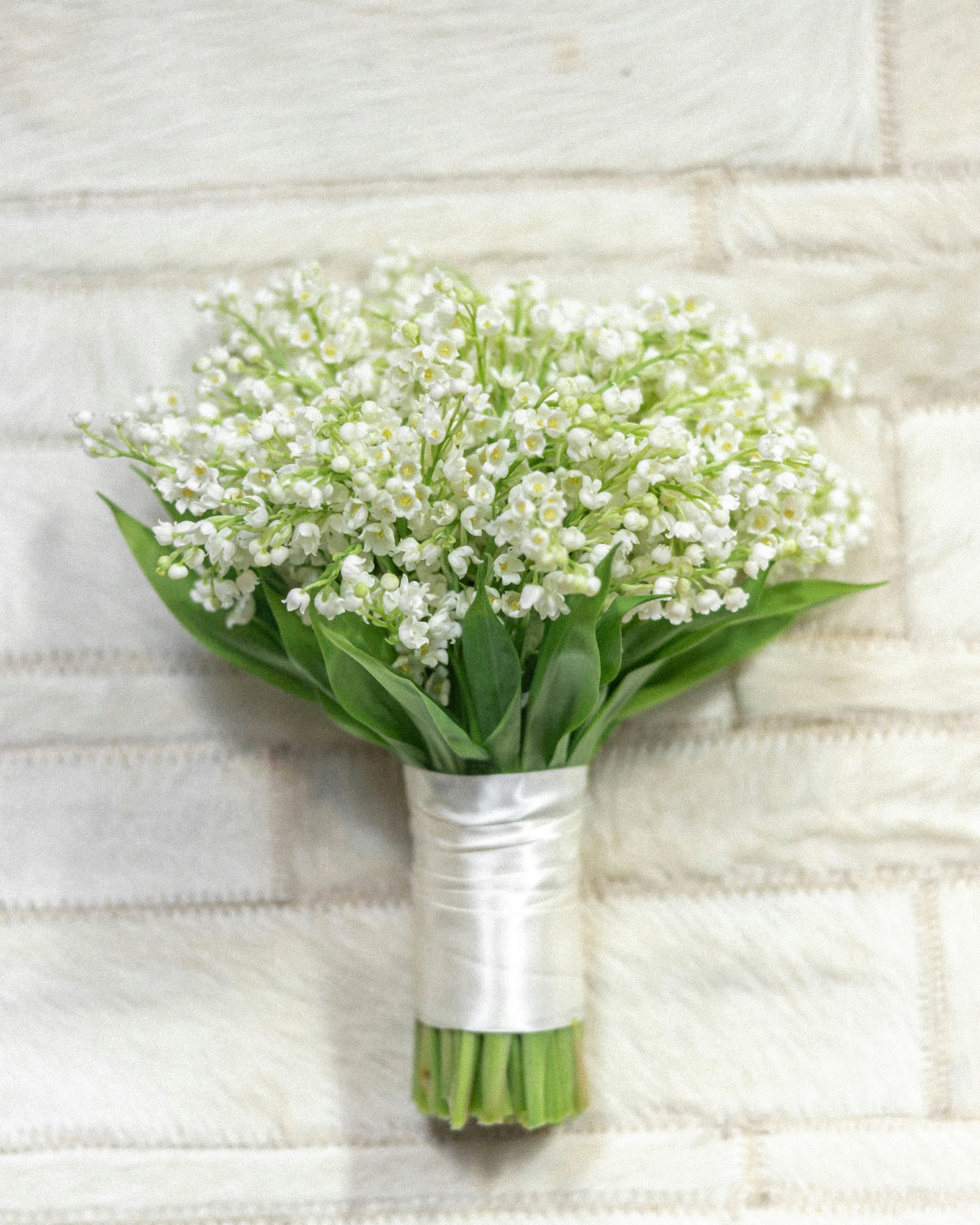 Bouquet of white lily of the valley flowers with green leaves wrapped in white ribbon.