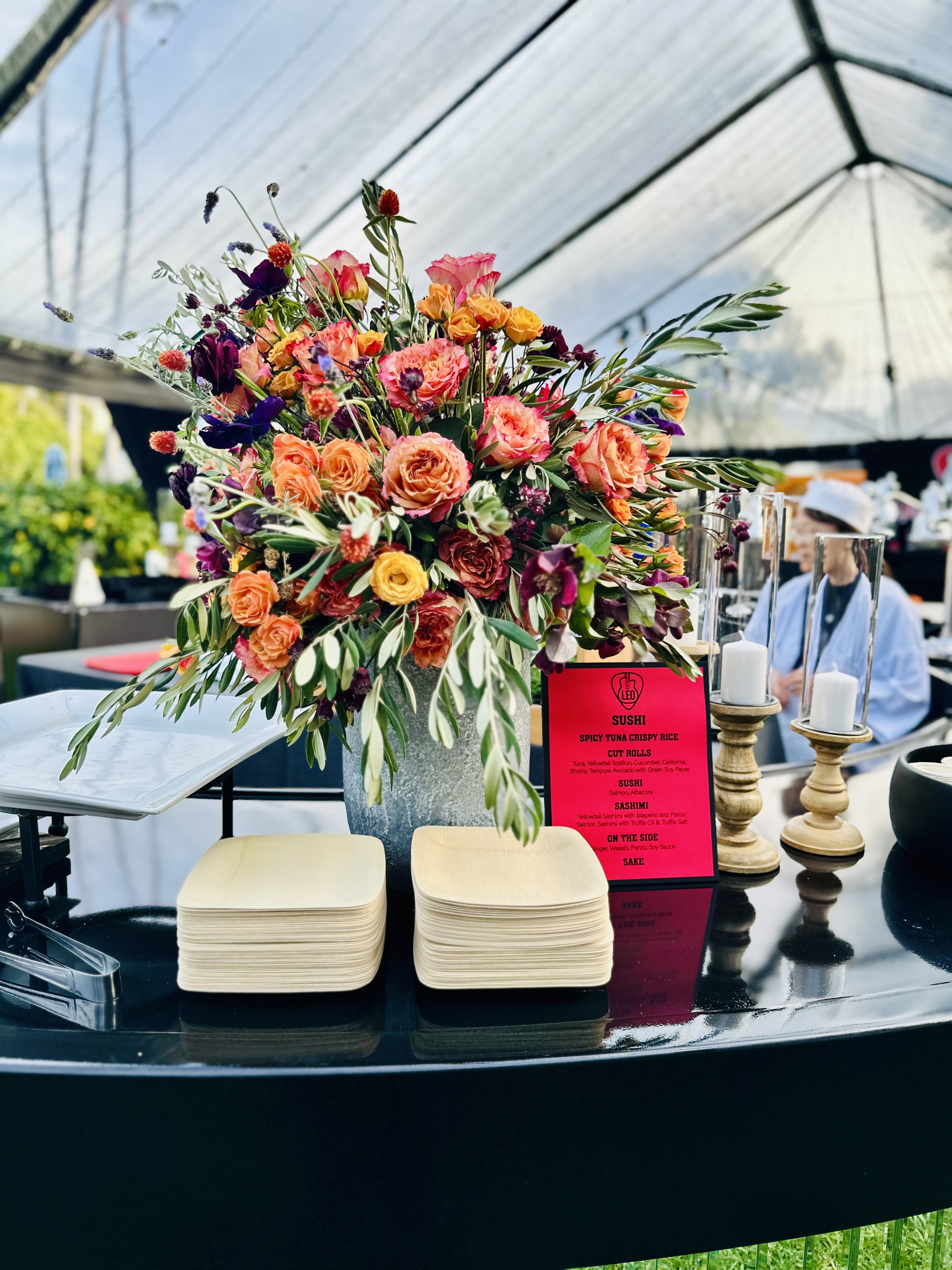 Large floral arrangement with orange, red, purple, and yellow flowers on a black table alongside stacked wooden plates, a bright red sushi menu, and two candles in glass holders under a tent.