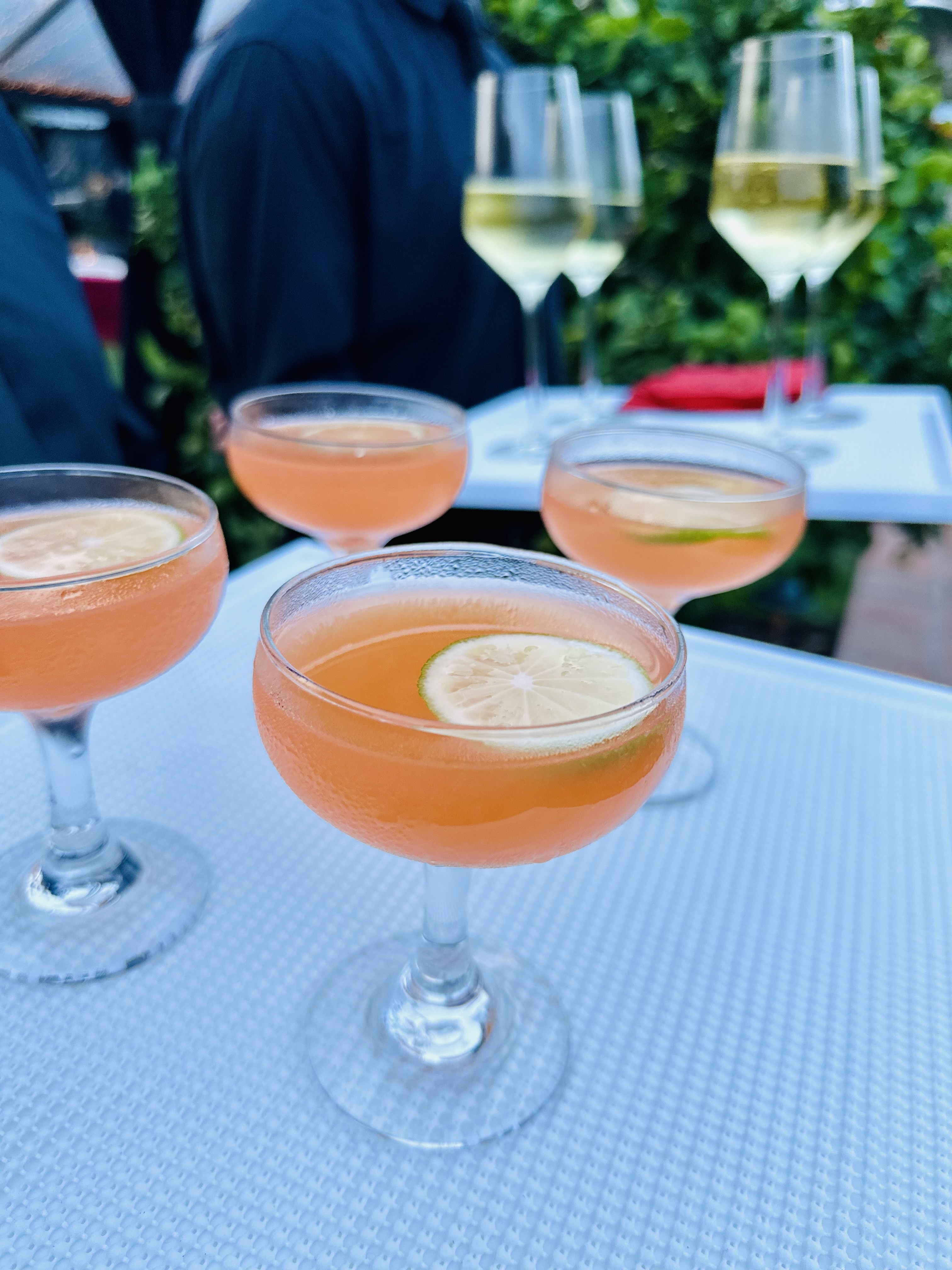 Four pink cocktails garnished with lime slices on a white textured table, with white wine glasses on trays held by servers in the background.