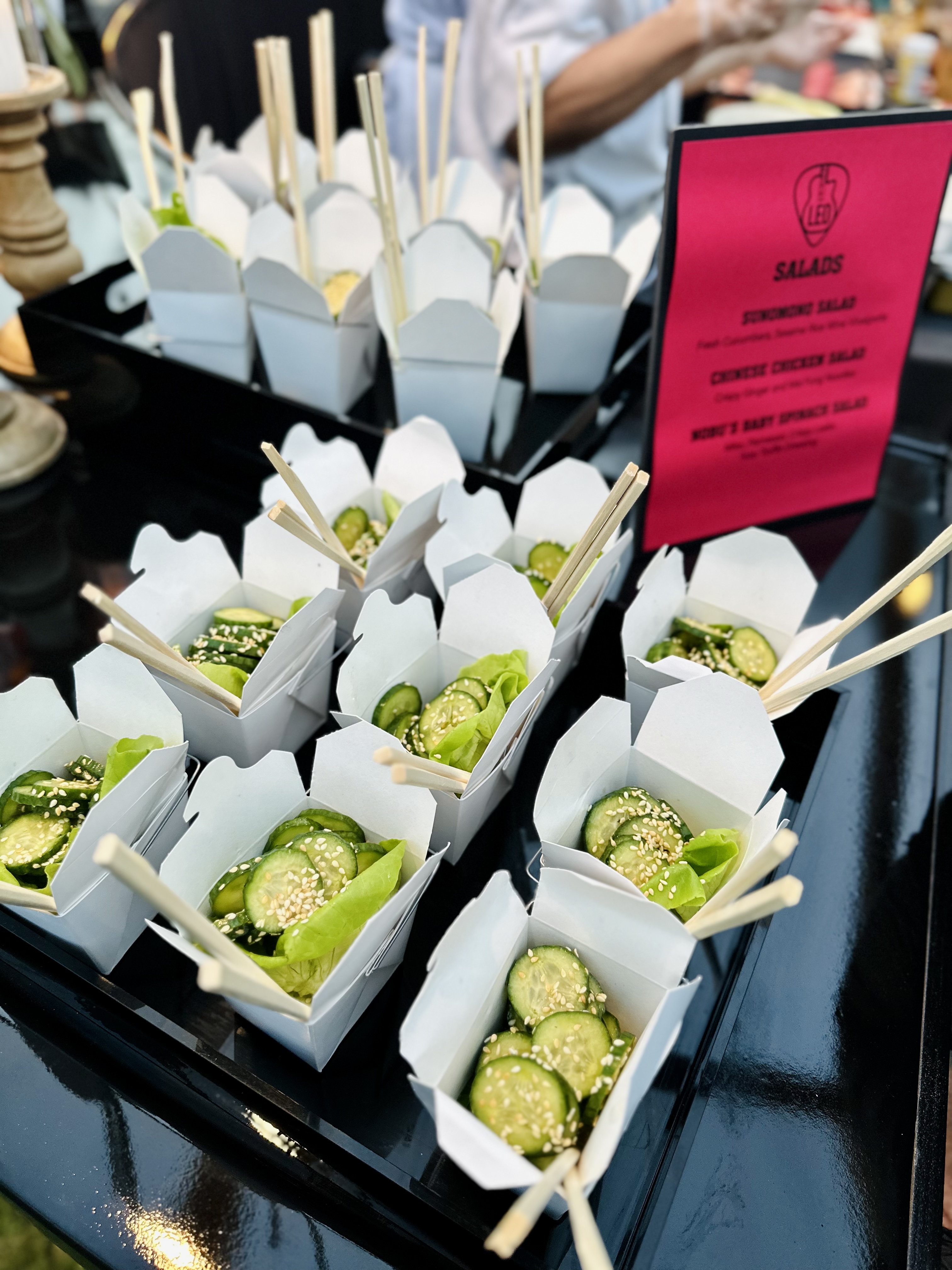 Small white takeout boxes filled with cucumber salad topped with sesame seeds and lettuce, each with chopsticks inserted.