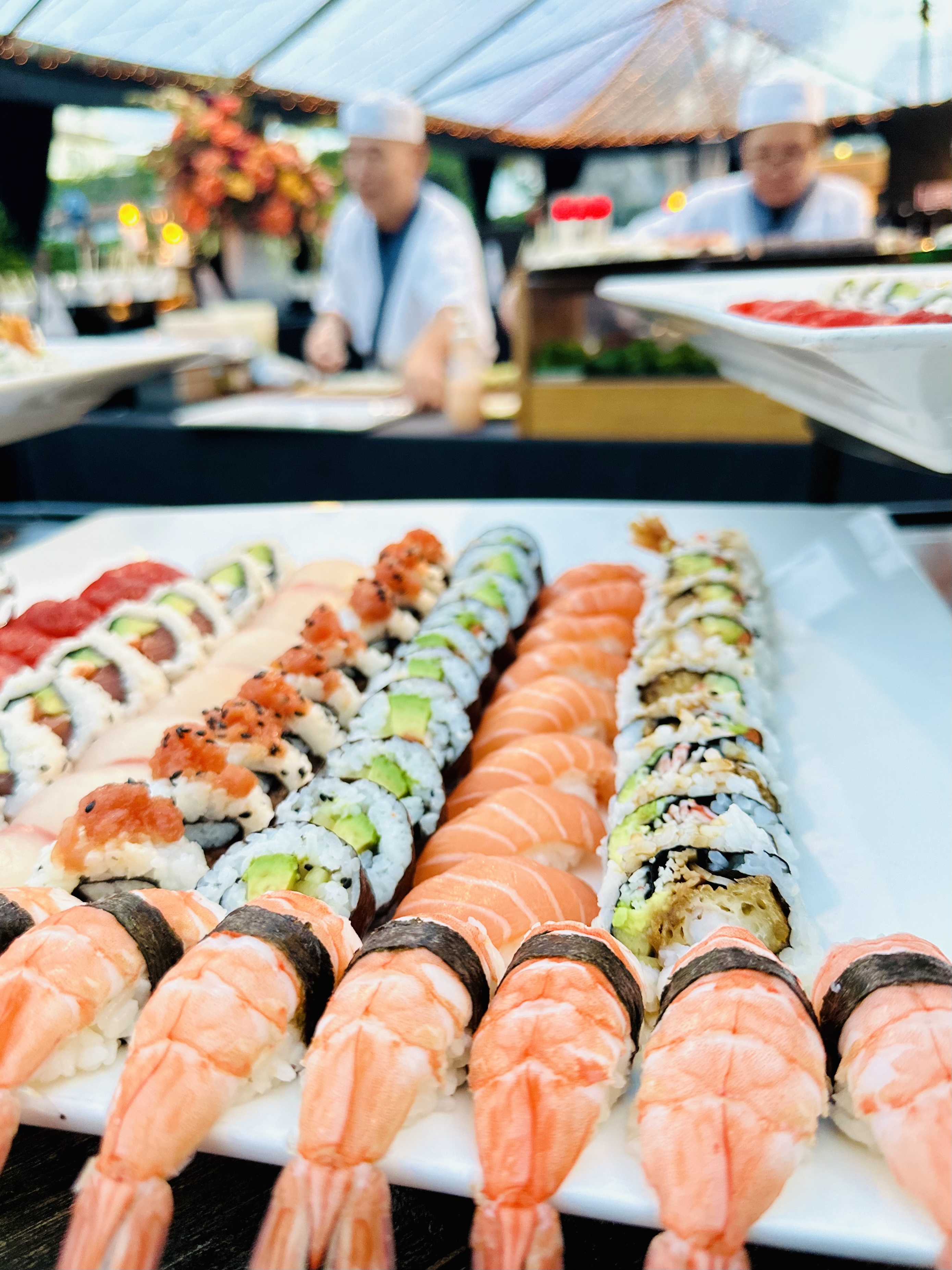 Close-up of a large plate of assorted sushi rolls and nigiri with chefs blurred in the background.