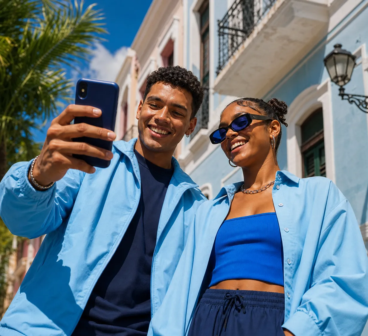 Dos jóvenes sonrientes toman una selfie al aire libre frente a un edificio azul bajo el sol.