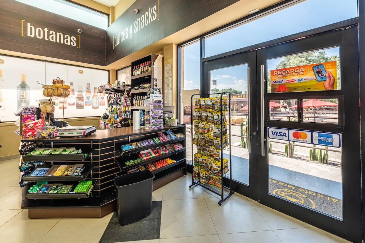 Interior de tienda con mostrador de snacks, botanas y dulces junto a puerta de entrada con cartel de recarga y tarjetas de crédito.