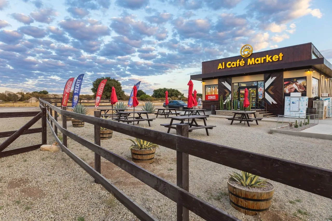 Mercado Al Café con mesas de picnic y sombrillas rojas al aire libre, rodeado de un paisaje rural y cielo parcialmente nublado.
