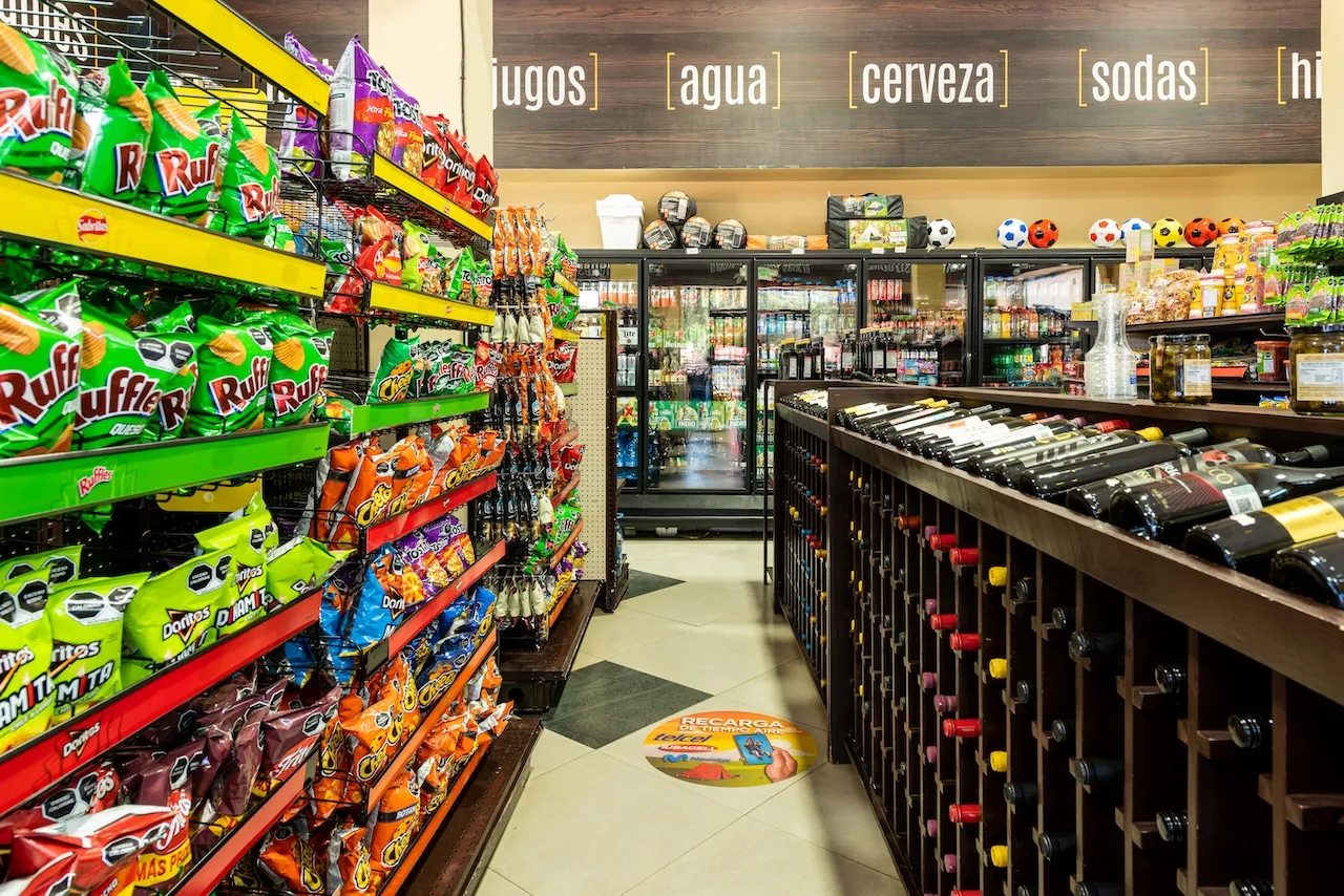 Pasillo de tienda con estantes llenos de botanas y refrescos a la izquierda, y botellas de vino en exhibidor de madera a la derecha.