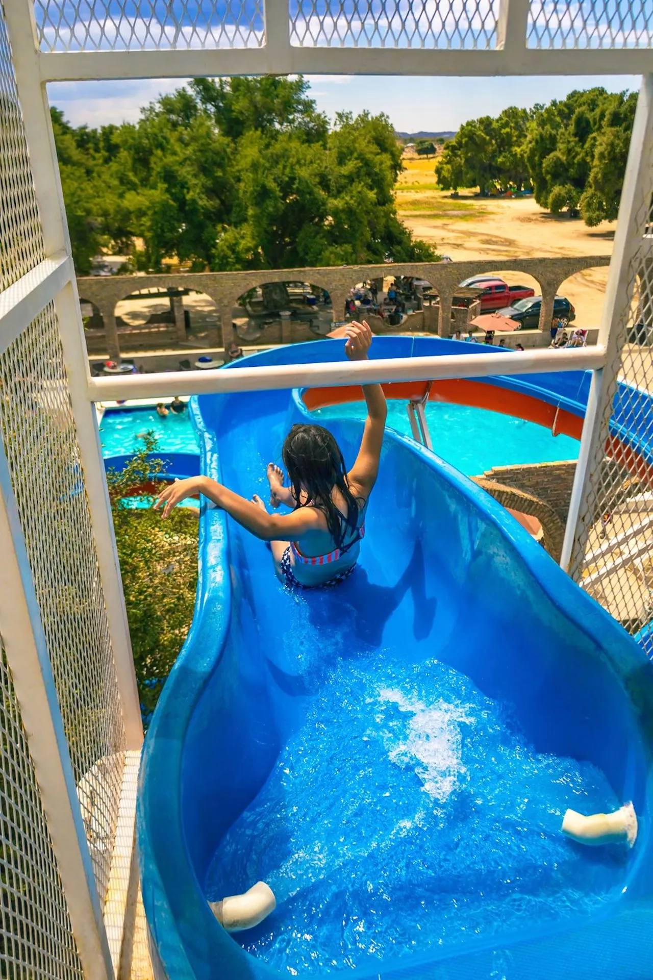 Niña bajando por un tobogán azul de parque acuático con árboles y vehículos en el fondo.
