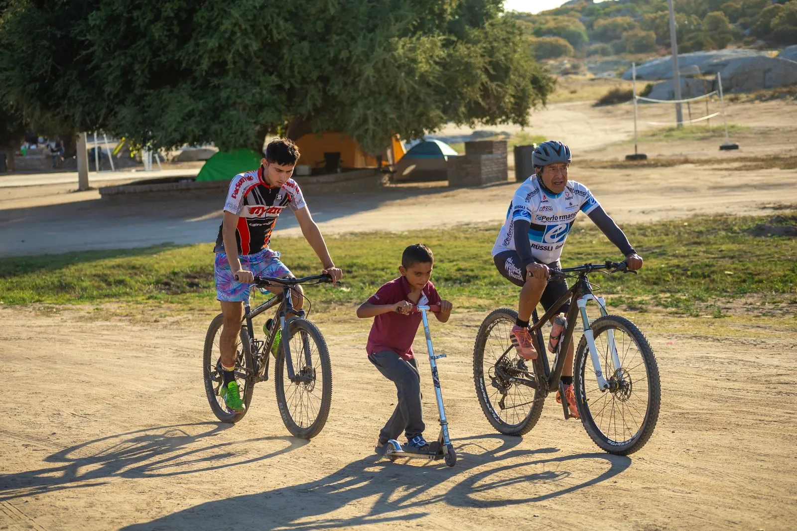 Dos hombres en bicicletas de montaña y un niño en scooter disfrutando al aire libre en un camino de tierra.