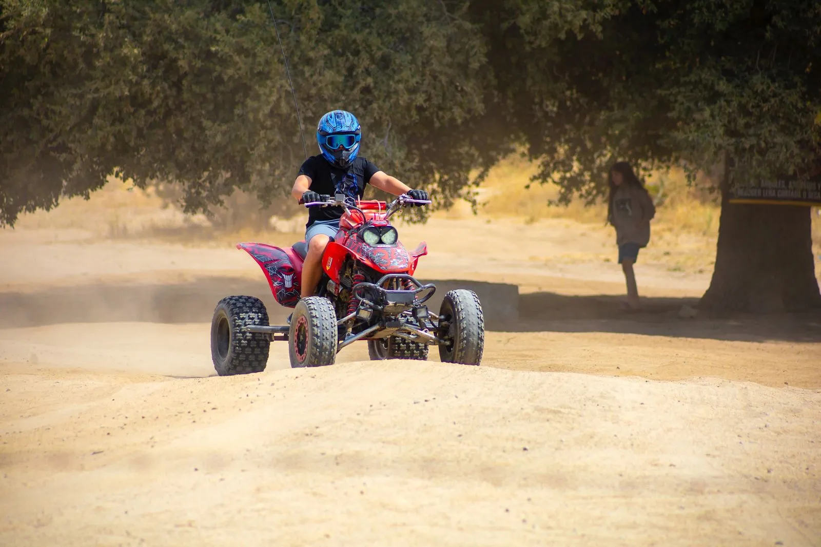 Persona con casco azul conduciendo un cuatrimoto rojo en un camino de tierra con árboles al fondo.
