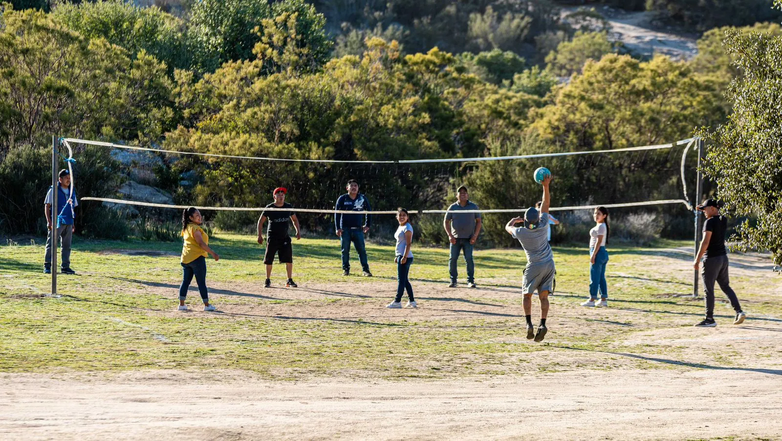Grupo de personas jugando voleibol al aire libre en una cancha rodeada de árboles.