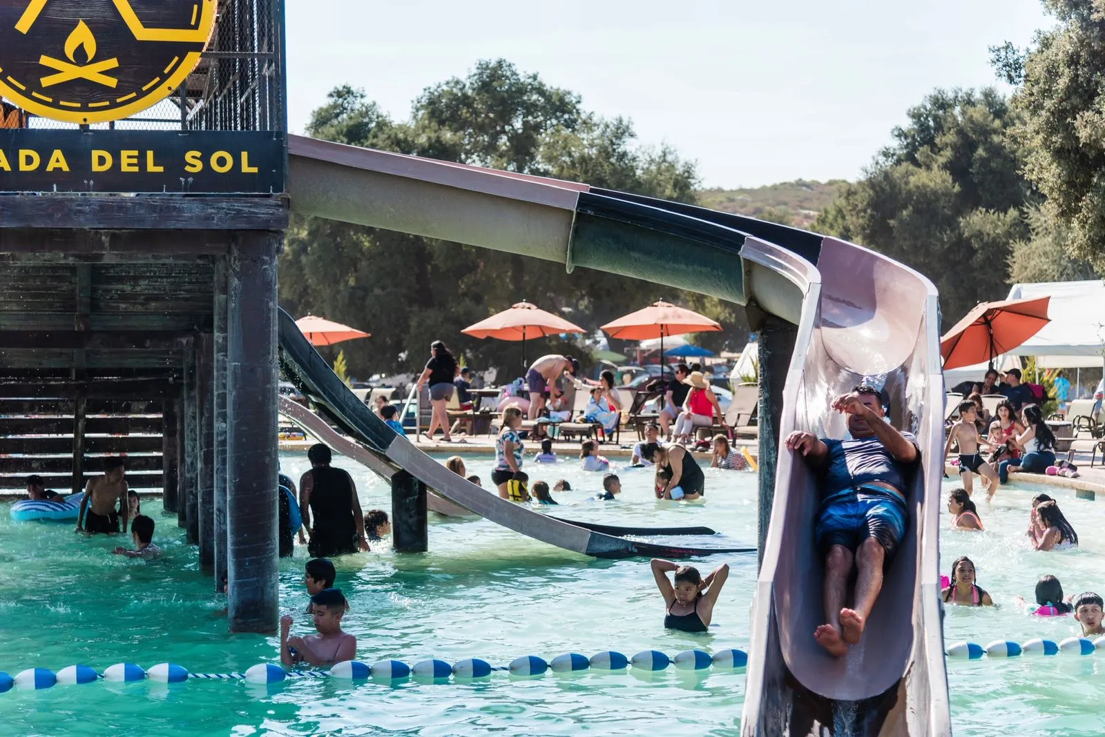 Persona bajando por un tobogán en una piscina llena de gente disfrutando y sombrillas naranjas al fondo.
