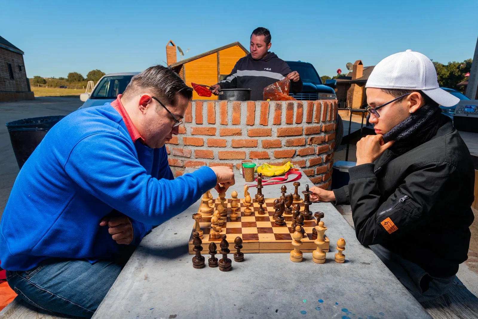 Dos hombres juegan ajedrez en una mesa al aire libre mientras otro hombre cocina junto a una estructura de ladrillo.