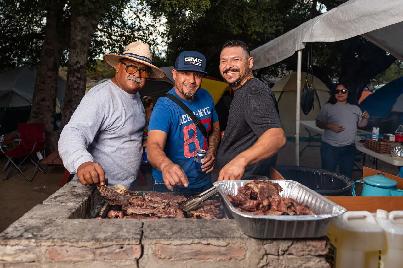 Tres hombres sonrientes cocinando carne en una parrilla de ladrillo durante un camping al aire libre con tiendas de campaña al fondo.
