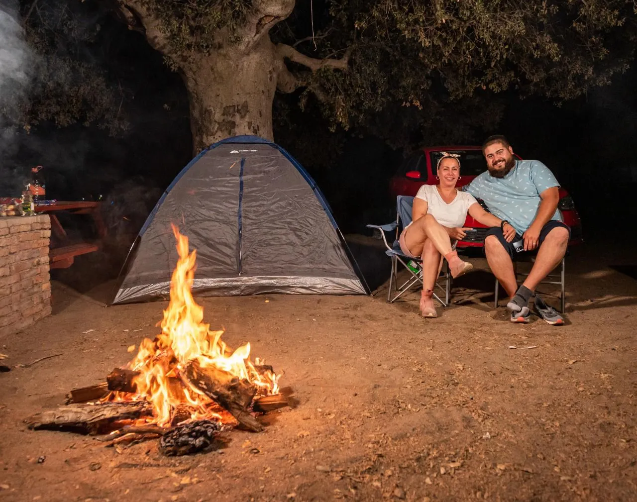 Pareja sonriente sentada en sillas de camping cerca de una fogata, una tienda y un coche rojo de noche.
