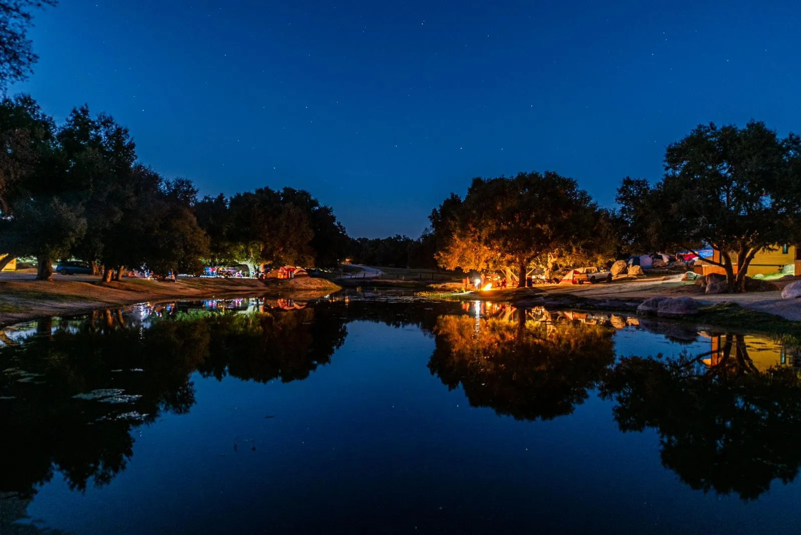 Escena nocturna de un campamento junto a un lago con árboles y fogata reflejados en el agua bajo un cielo estrellado.