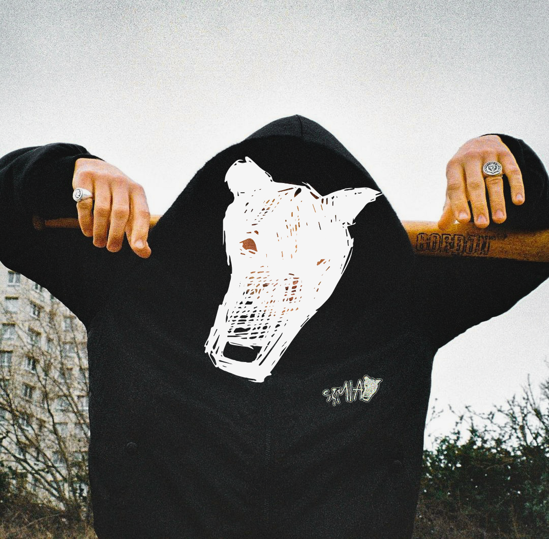 Person wearing a black hoodie with a white abstract animal face on the hoodie, holding a wooden baseball bat behind their neck, showing rings and a tattoo on the arm.