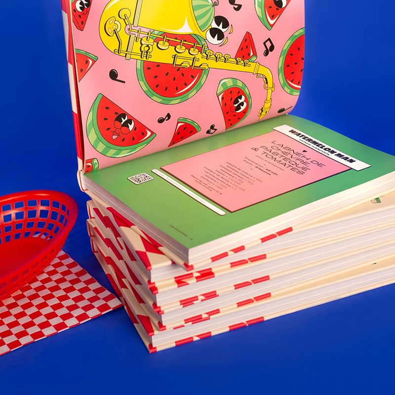Stack of books with red and white checkered spines, one open showing a pink page with cartoon watermelons and a yellow saxophone illustration on a blue background.