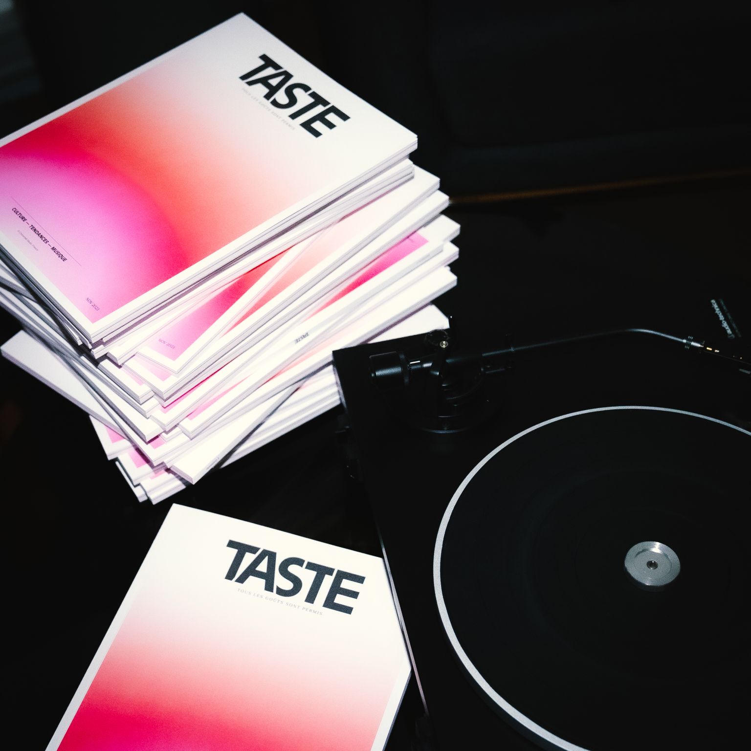 Stack of magazines titled 'TASTE' with a pink gradient cover placed next to a black turntable.