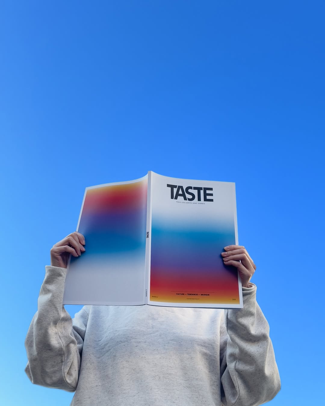 Person wearing a light gray sweatshirt holding and reading a magazine titled 'TASTE' against a clear blue sky.