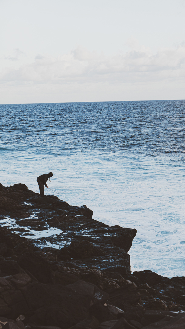 Person fishing on rocky coastline with ocean waves in the background under a cloudy sky.