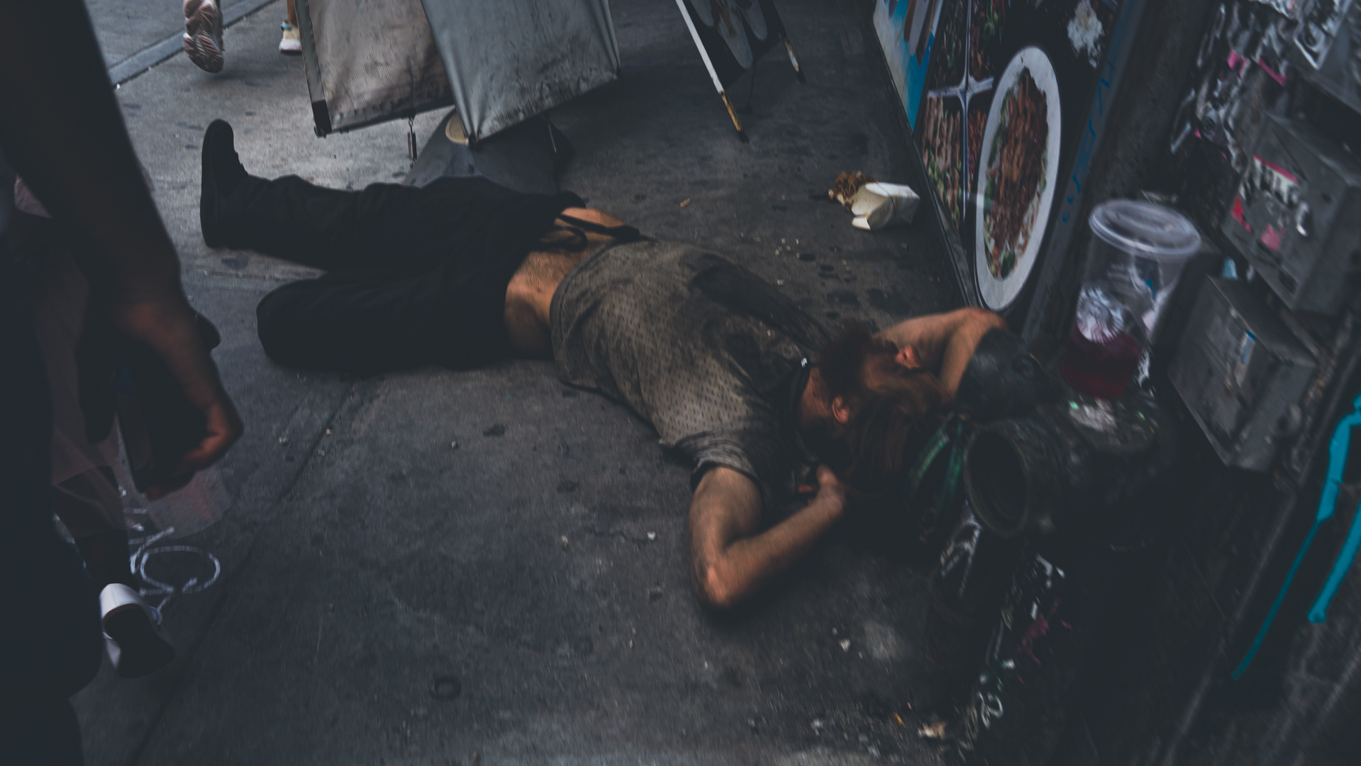Person lying on a dirty sidewalk next to a fire hydrant and a food vendor’s wall with food pictures.