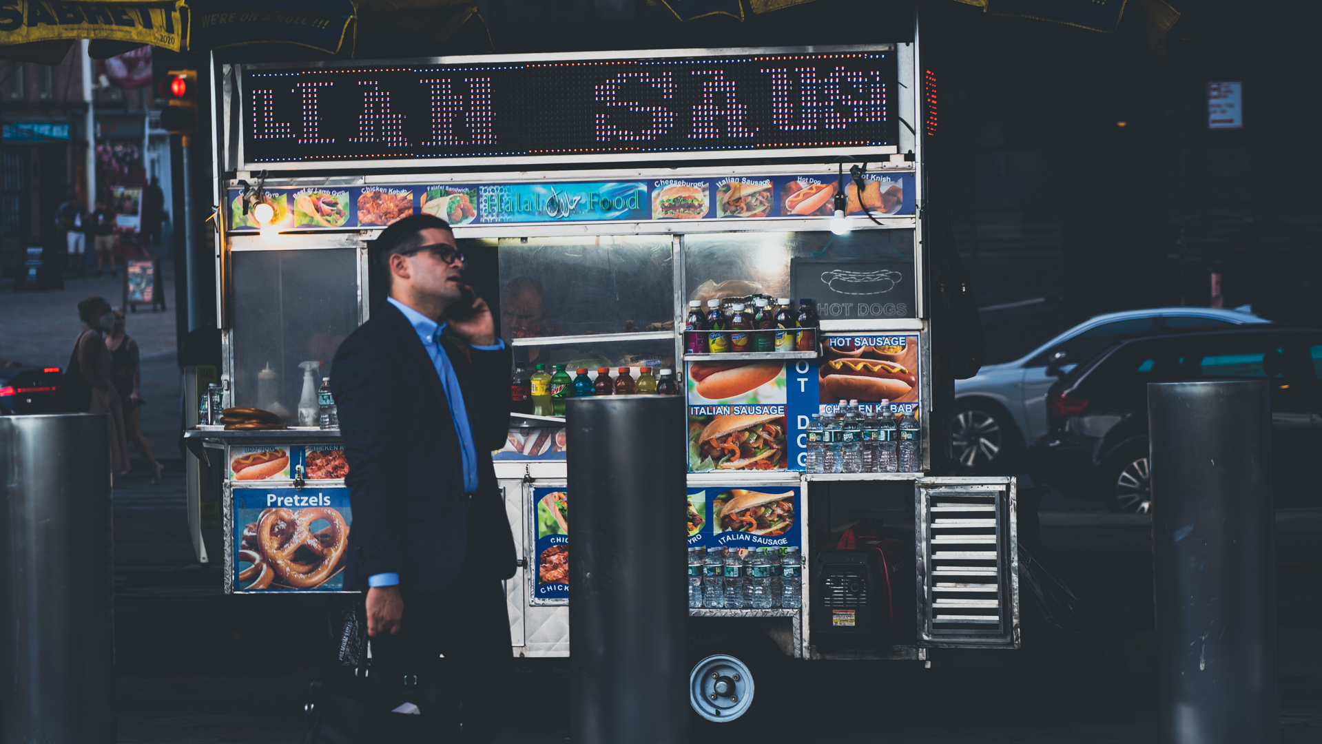 Man in a suit talking on a phone walking past a street food cart selling hot dogs, Italian sausage, pretzels, and bottled drinks at dusk.