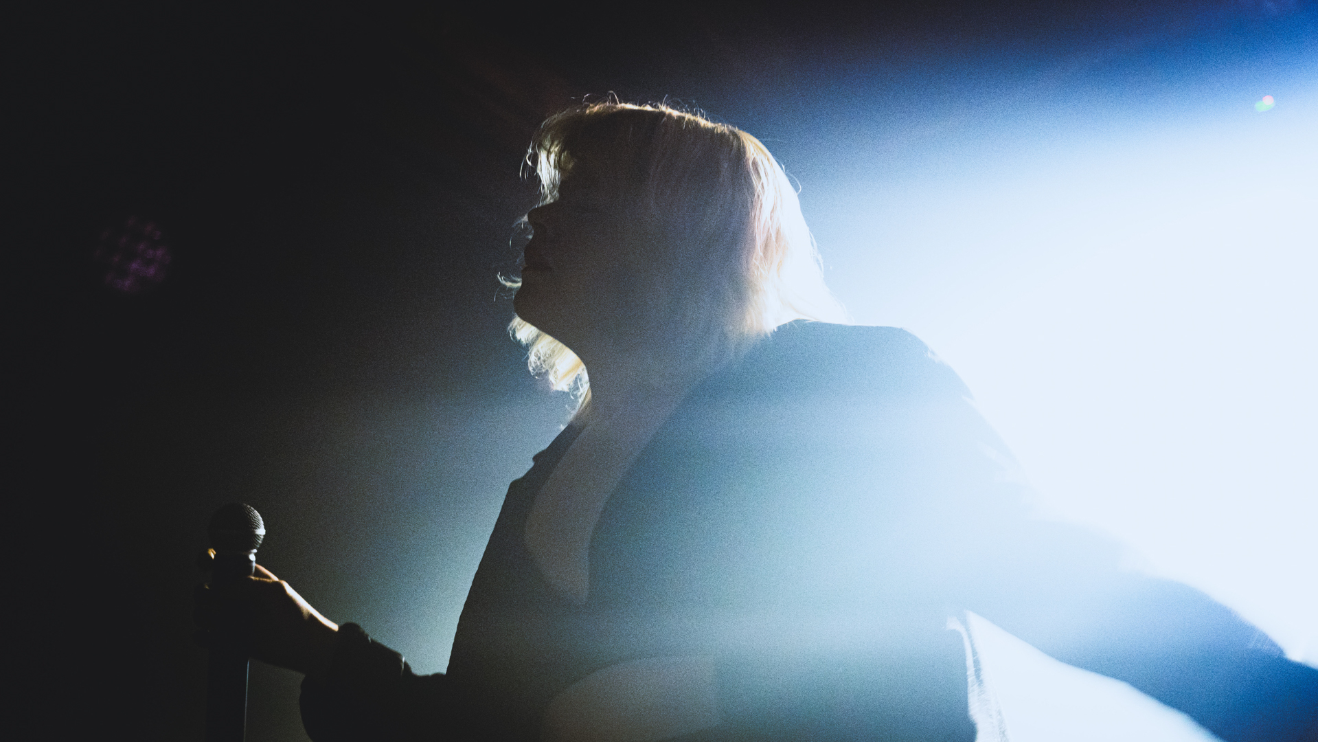 Silhouette of a person with light-colored hair holding a microphone, illuminated by bright backlight on a dark stage.