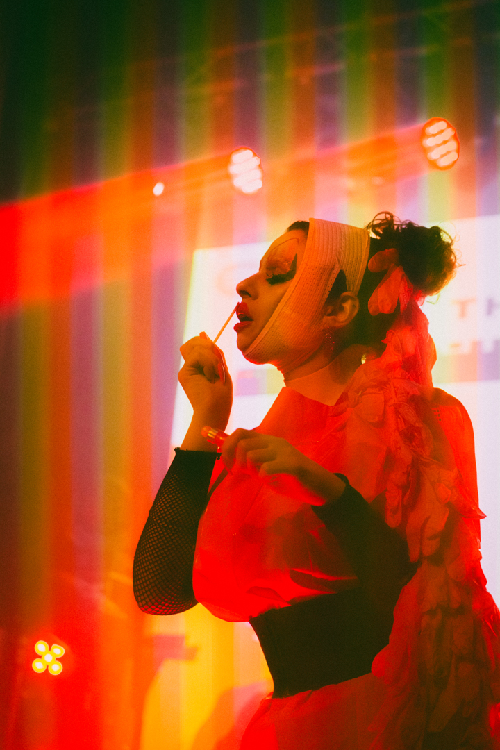 Person wearing dramatic makeup and a red costume applying red lipstick under warm stage lighting.