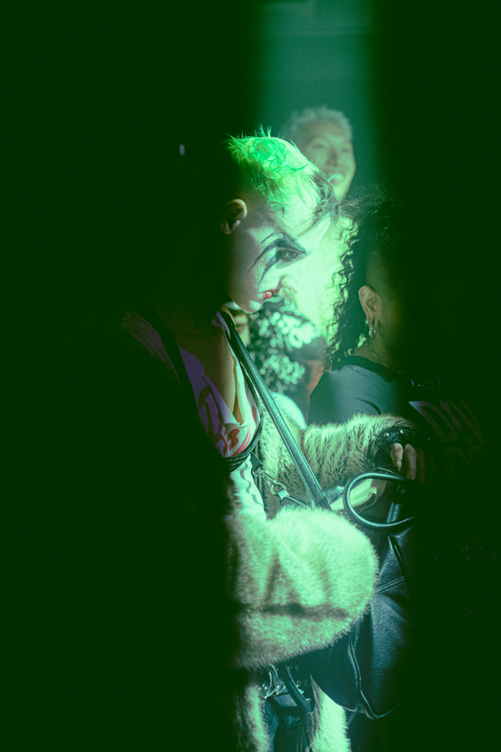Person with dramatic face makeup and green hair, illuminated by green light in a dark setting, wearing a fur coat and holding a bag.