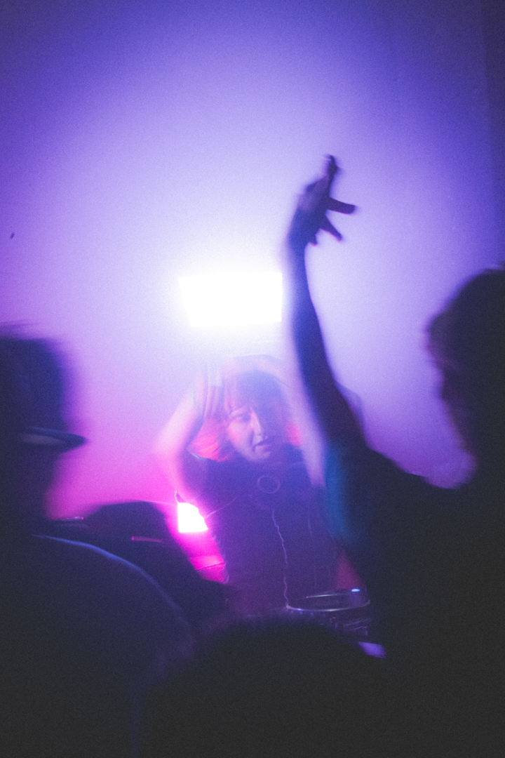 Person with headphones DJing under purple and pink lighting, with blurred hands and crowd in foreground.