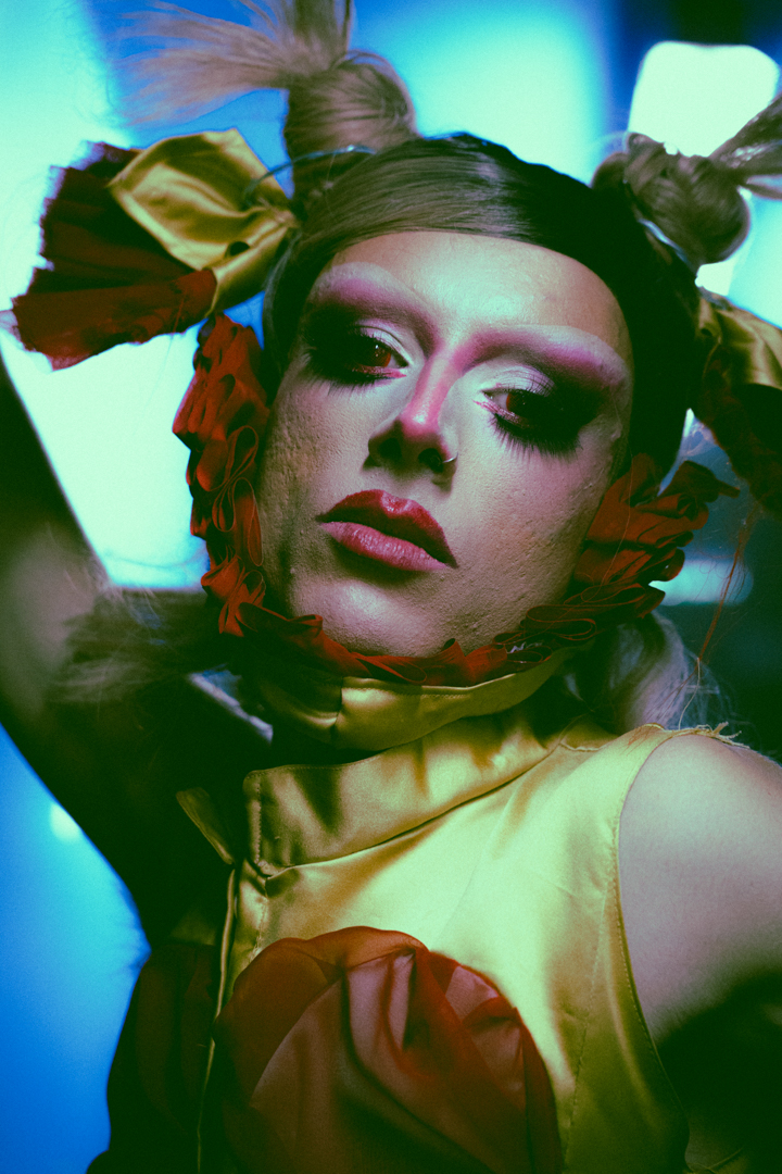 Close-up portrait of a person with dramatic makeup, wearing a yellow outfit and styled hair with ribbons.