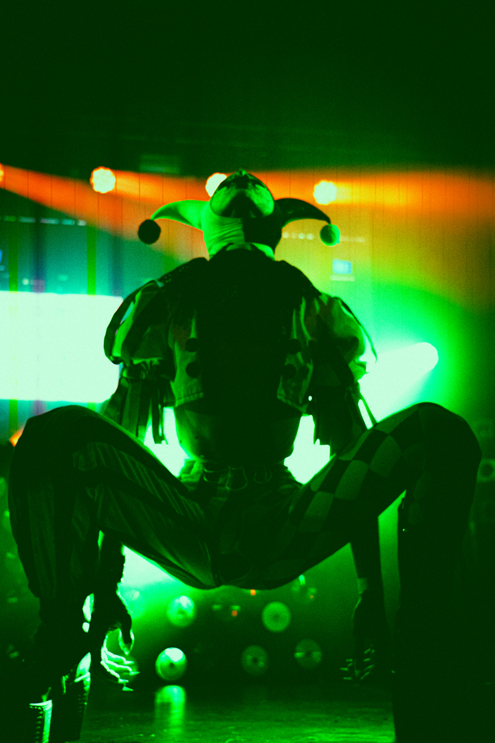 Performer in a jester costume with checkered pants striking a low, wide pose on stage under green and orange lighting.